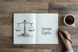 Open book showing hand sketching scales of justice and words reading Human Rights resting on a table next to a coffee