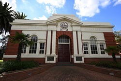 The front entrance of the historic red brick City of Oakleigh Council Chambers. The building features a white classical portico with columns and a pediment. Above the entrance, there is a circular emblem with the date “1921” inscribed. The architecture reflects early 20th-century design. The entrance is framed by palm trees, and a clear blue sky serves as the backdrop