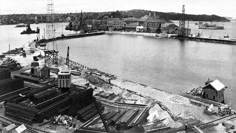 An old black and white photo of a harbor with construction equipment in the foreground.